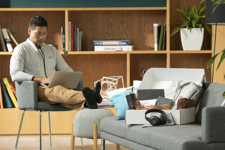 A new hire gift box including company-branded headphones, tumbler, hoodie, notebook, and welcome card, next to an employee wearing an AVEVA branded quarter-zip.