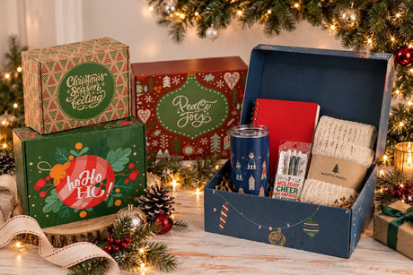A festive collection of corporate holiday gift boxes, including an open box with useful everyday gifts such as a tumbler, notebook, blanket, and stationery, styled for employee and client holiday gifting.