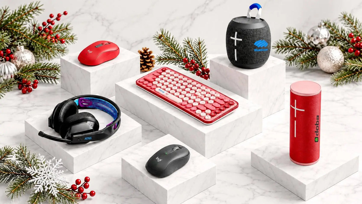 Branded tech gifts including wireless mice, a red keyboard, a gaming headset and portable Bluetooth speakers displayed on white marble blocks with pine branches, red berries and silver ornaments.
